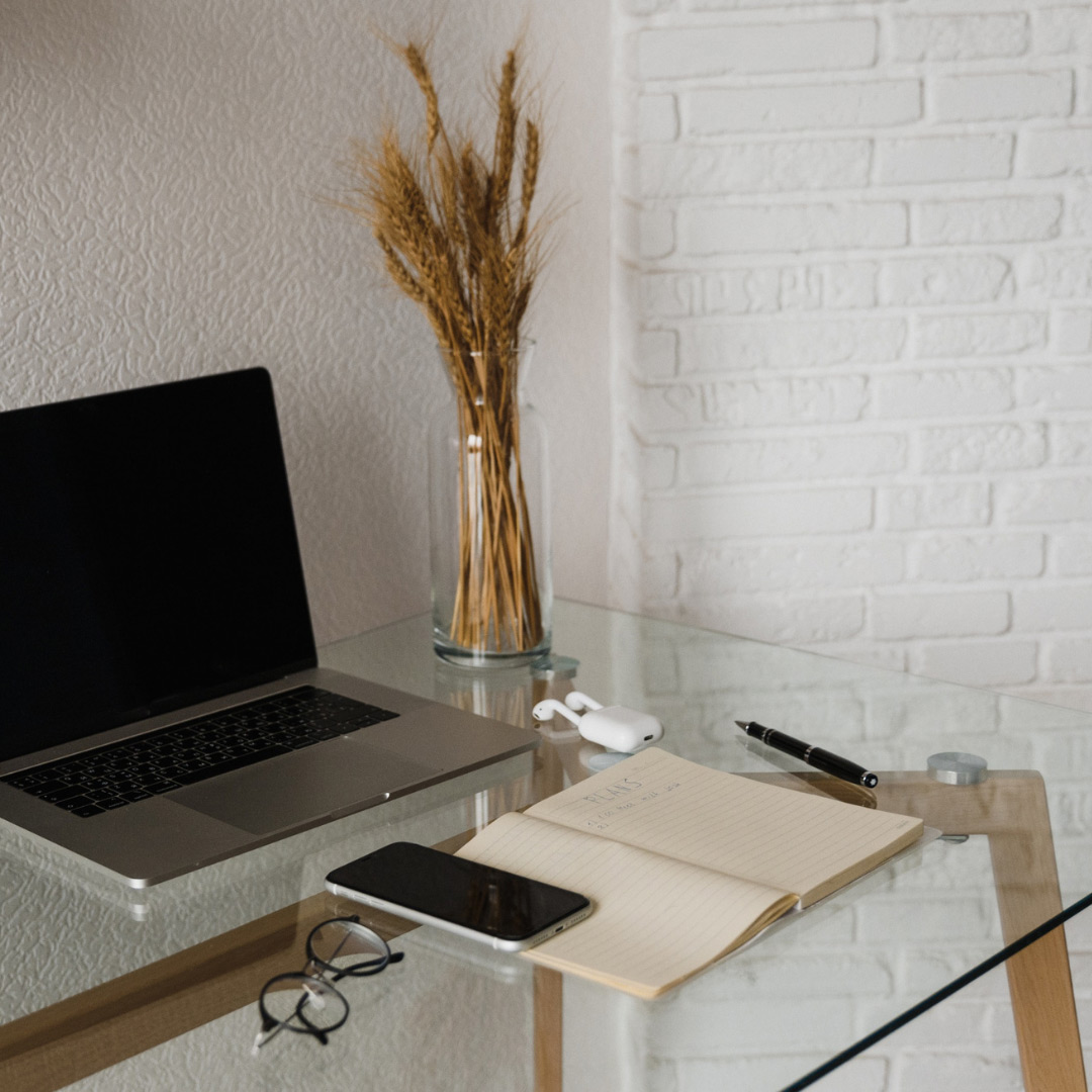 Laptop on glass desk with notepad and phone.